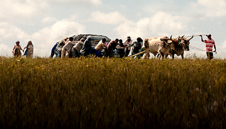 Image from Propriedade; a group of people, and two oxen, tow a tightly bound car across a wheat field.